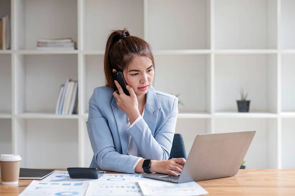 Attractive Asian woman using laptop in online meeting. Businesswoman using mobile phone talking with colleague sitting at table in modern office. - Photo, Image