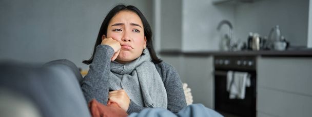 Obraz smutnej młodej kobiety źle się czującej, siedzącej z szalikiem i marszczącej brwi w salonie, przeziębionej i przebywającej w domu w izolacji. - Zdjęcie, obraz