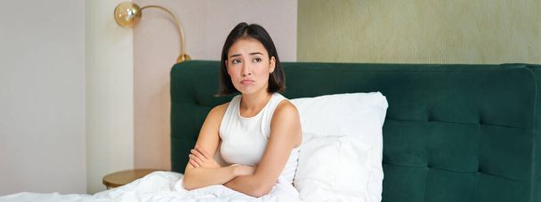 Portrét asijského děvčete v posteli, vypadá komplikovaně a ustaraně, zkřížené paže na hrudi, zamračený neklidným, přemýšlivým výrazem ve tváři. - Fotografie, Obrázek