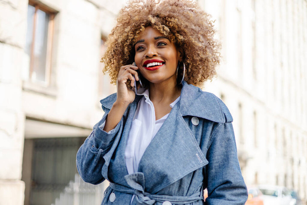 Beautiful black woman walks down the street and uses smart phone for communication. She is happy and smiled. Bright sunny day. - Φωτογραφία, εικόνα