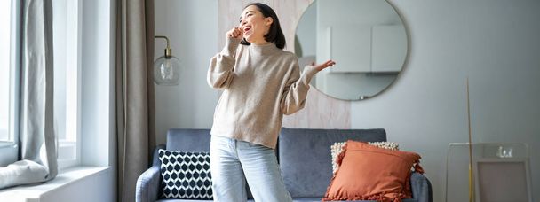 Personas y emociones. Retrato de chica asiática de pie en la sala de estar, cantando en el micrófono de sombra, bailando y divirtiéndose, sintiendo alegría. - Foto, Imagen