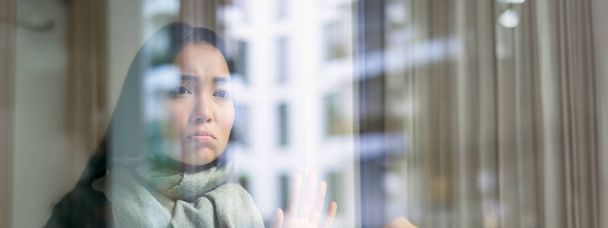 Asiatin fühlt sich unwohl, bleibt zu Hause, blickt mit eifersüchtigem, verzweifeltem Gesicht aus dem Fenster, will nach draußen, wird aber kalt erwischt. - Foto, Bild