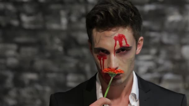 男性の吸血鬼は花の匂いを嗅ぎ、それを与えます。花に焦点を当てたラック。レンガの壁の黒い背景にポーズ吸血鬼のイメージの男。ハロウィーンのコンセプトと衣装 - 映像、動画
