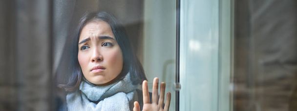 Vertikale Aufnahme einer aufgebrachten asiatischen Frau, die nach draußen schaut, Fenster berührt, mit düsterem eifersüchtigen Gesicht auf die Straße starrt, sich krank fühlt, mit Koliken oder Grippe zu Hause bleibt. - Foto, Bild
