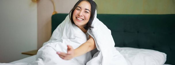 Женщина сидит на кровати, накрытая белым одеялом, улыбается, радуется счастливому уик-энду, смеется перед камерой. - Фото, изображение