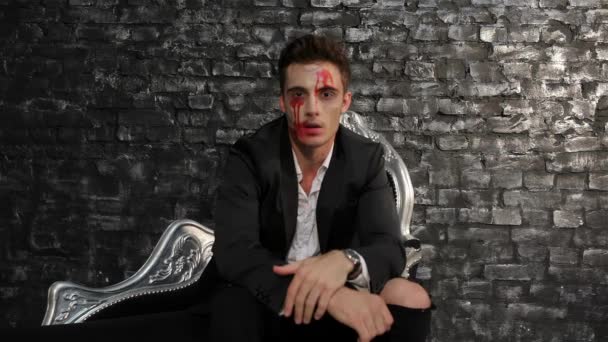 Male vampire sitting on a black sofa. Man in the image of the vampire posing on black background of a brick wall. Halloween concepts and costumes - Footage, Video