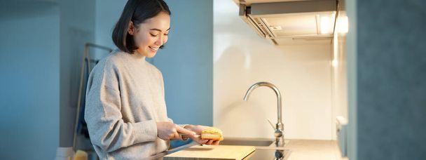 Portrait de sourire jeune femme asiatique debout sur la cuisine et faire un sandwitch, cuisine pour elle-même. - Photo, image