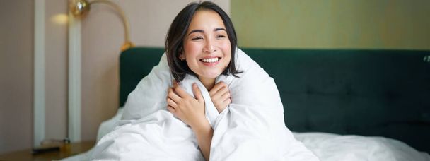 暖かいデュベットのベッドに座っている可愛くて柔らかい女性は、窓の外を見て笑って、朝を楽しんで毛布の快適さ. - 写真・画像