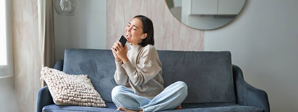 Portrait de fille asiatique insouciante est assis à la maison, écoute de la musique dans les écouteurs et chante sur smartphone, tient le téléphone mobile comme un microphone. - Photo, image