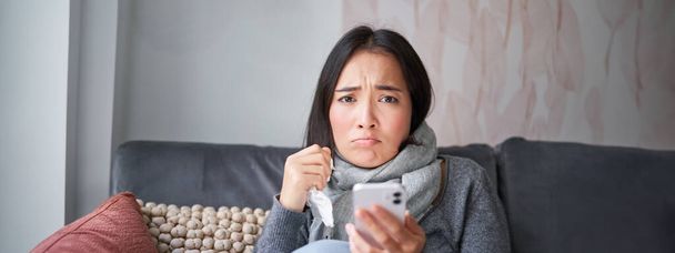 Aziatisch meisje met influenza, zit thuis, huilt en kijkt boos, houdt smartphone, voelt zich onwel en somber, het dragen van warme kleren. - Foto, afbeelding
