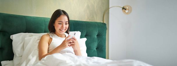 Piękny uśmiech azjatki kobieta w łóżku, czytanie wiadomości na smartfonie i śmiech, korzystając leniwy dzień w sypialni, za pomocą telefonu komórkowego. - Zdjęcie, obraz