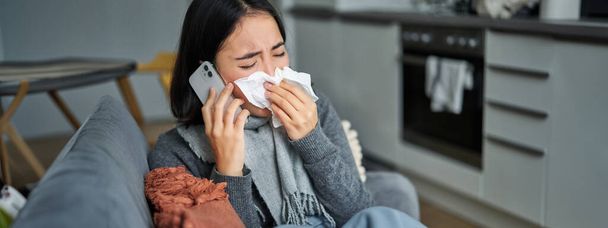 Portret van een zieke jonge Koreaanse vrouw die zich ziek voelt, niest en servet vasthoudt, ziek thuis blijft, verkouden is. Praten op mobiele telefoon. - Foto, afbeelding