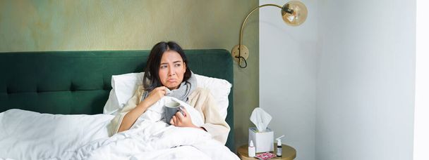 Covid-19, griep en vaccinatie concept. Koreaanse meisje liggend in bed met koude, griep vangen, het drinken van warme thee en met behulp van voorgeschreven medicatie. - Foto, afbeelding