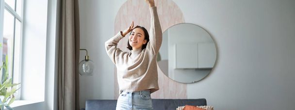 Naiset ja hyvinvointi. Kaunis nuori aasialainen nainen tanssii, tuntee olonsa huolettomaksi ja onnelliseksi, nostaa kätensä pään yläpuolelle ja hymyilee, nauttien olostaan kotona. - Valokuva, kuva