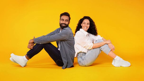 Happy millennial middle eastern spouses sitting on floor together, posing back to back on studio floor over yellow background, panorama. Great offer for young family couple concept - Zdjęcie, obraz