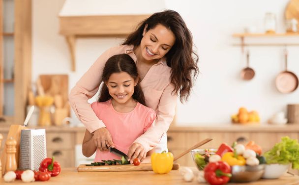 Glückliche Mutter bringt ihrem Kind das Kochen bei, schneidet Gemüse für den Salat zusammen und genießt die Zubereitung des Abendessens am Küchentisch im Haus. Familienernährung und gesunde Rezepte - Foto, Bild