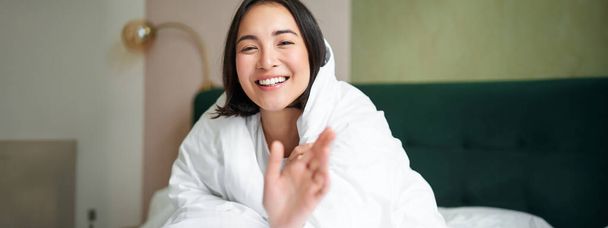 Happy beautiful asian woman covered with blanket, sits on bed in warm duvet, tries to reach camera with a hand, smiling playfully. Hotel bedroom, comfort and apartments concept. - Photo, Image