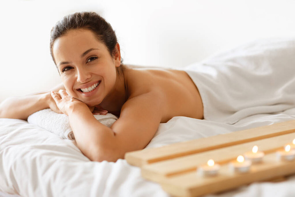 Attractive Brunette Young Woman Relaxing During Spa Treatment In Beauty Salon, Lying On Stomach On Massage Table Near Burning Candles, Smiling To Camera Indoor. Pampering And Wellness - Photo, Image