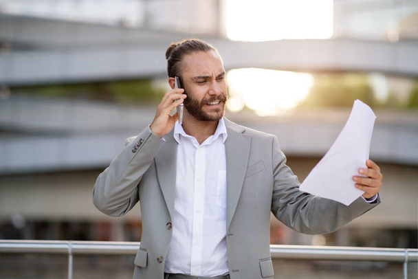 Business Problems. Angry Young Businessman Holding Papers And Talking On Cellphone Outdoors, Furious Male Entrepreneur Standing Outside Near Modern Office Building, Managing Issues By Phone - Photo, Image