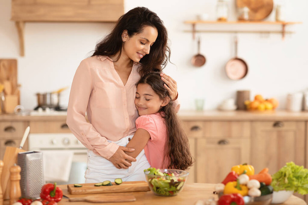 Liebevolle Mutter, die ihre Tochter umarmt, während sie gemeinsam in der modernen Küche kocht und Liebe und Sorgfalt für die Ernährung der Familie ausdrückt. Kind umarmt Mama bei Essenszubereitung zu Hause - Foto, Bild