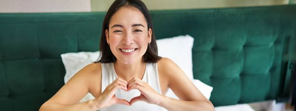 Linda chica asiática muestra el corazón, signo de amor, acostado en la cama y sonriendo, disfrutando de las mañanas, días perezosos en el dormitorio. - Foto, Imagen
