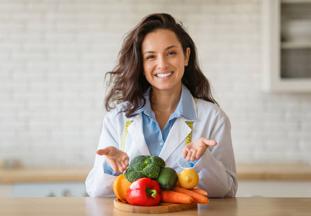 Portrait d'une diététiste joyeuse en blouse de laboratoire pointant vers les fruits et légumes frais sur la table, souriant à la caméra à la clinique. Perte de poids consultant recommandant une alimentation saine à base de plantes - Photo, image