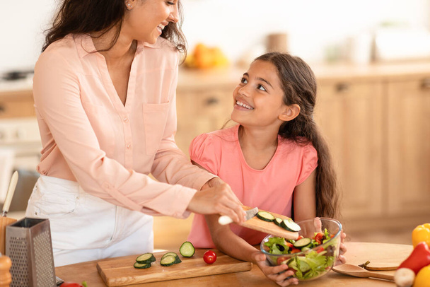 Jovem mãe e filha desfrutando da preparação do jantar juntos, cozinhando salada com legumes orgânicos frescos em casa, adicionando ingredientes para tigela, de pé perto da mesa da cozinha interior. Cultivadas - Foto, Imagem
