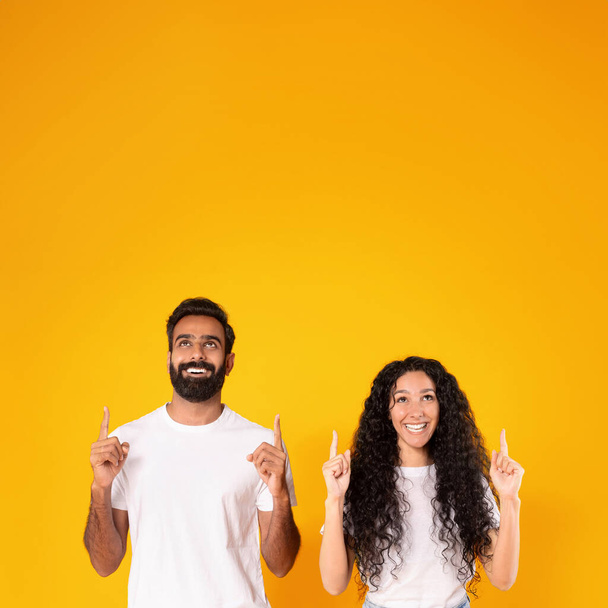 Mira esto. Foto cuadrada de feliz pareja árabe apuntando con los dedos hacia arriba con ambas manos sobre el fondo amarillo del estudio. Jóvenes cónyuges mostrando el espacio vacío hacia arriba sobre sus cabezas. Gran oferta - Foto, Imagen