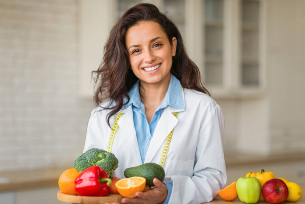 Mosolygó női táplálkozási szakember portréja egy tányér friss gyümölccsel, aki a fogyókúrás klinikán dolgozik, pózol és mosolyog a kamerába. Egészséges táplálkozási tanácsadó egészséges táplálkozást javasol - Fotó, kép