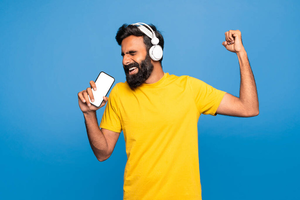 Hombre indio feliz en auriculares inalámbricos cantando en el teléfono inteligente con pantalla vacía en micrófono imaginario, de pie sobre fondo azul. Karaoke para superestrella, música en tiempo libre - Foto, Imagen