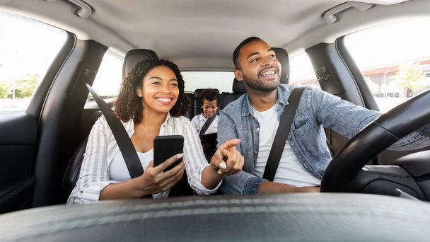 Pozytywna wesoła czarna rodzina ma przejażdżkę samochodem, korzystając ze smartfona, sprawdzając nawigację mapy podczas podróży. Szczęśliwy afrykański matka punkt na drodze, ojciec jazdy samochodem, dziecko siedzi z tyłu siedzieć - Zdjęcie, obraz