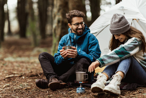 Positive young caucasian family in jackets sitting, relaxing near tent, prepare hot drink, enjoy adventure, free time at weekend in cold forest in autumn. Vacation together, relationship, lifestyle - Photo, Image