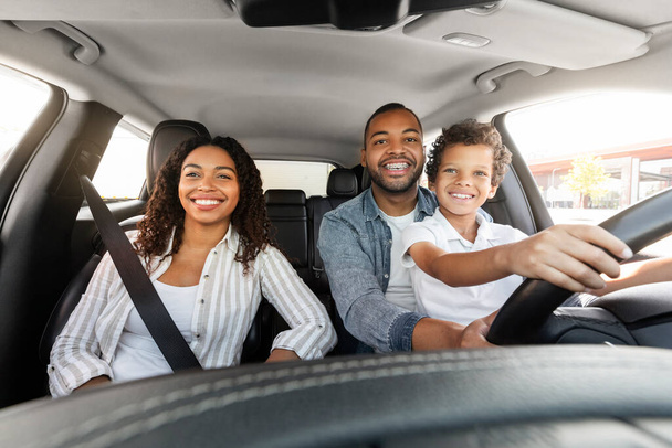 Glückliche schwarze Millennial-Eltern Vater und Mutter genießen nagelneues Auto mit ihrem kleinen Sohn. Aufgeregt preteen afrikanisch-amerikanische Junge sitzt auf Papa Schoß, Auto fahren zusammen und lächelt - Foto, Bild