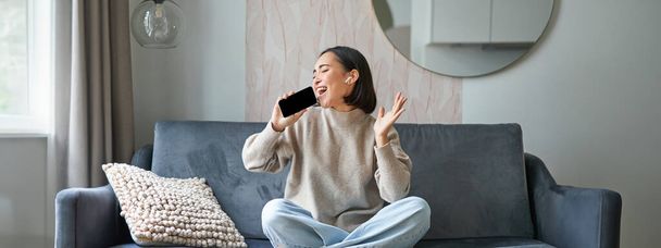 Портрет беззаботной азиатской девушки сидит дома, слушает музыку в наушниках и поет на смартфоне, держит мобильный телефон, как микрофон. - Фото, изображение