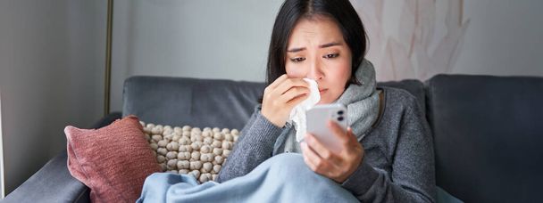 Portret van een zieke vrouw die contact opneemt met haar huisarts via mobiele telefoon terwijl ze verkouden is, thuis zit met griep, met een smartphone. - Foto, afbeelding