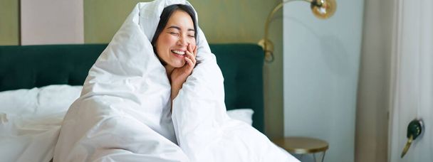 Krásná asijská žena sedí na posteli, přikrytá přikrývkami a přikrývkou, směje se a usmívá, užívá si víkend v ložnici. - Fotografie, Obrázek