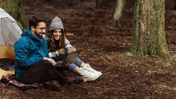 Positive young caucasian family in jackets sit near tent, chatting on laptop, enjoy adventure, weekend, travel blog in cold forest in autumn. Work, relationship, vacation together, active lifestyle - Photo, Image