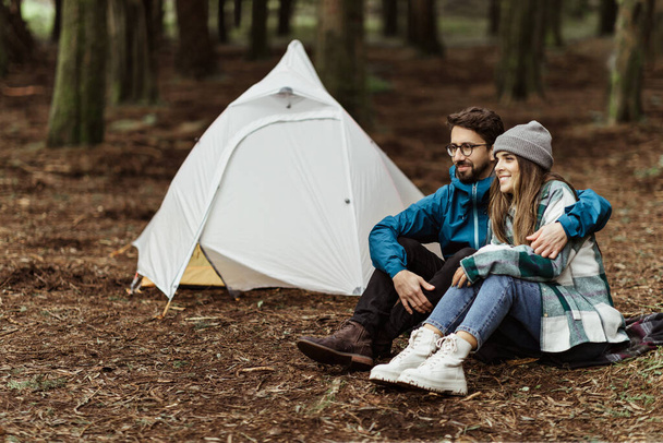 Happy young caucasian family in jackets sitting, relaxing near tent, enjoy adventure, free time at weekend in cold forest in autumn. Love, relationship, vacation together, active lifestyle - Photo, Image