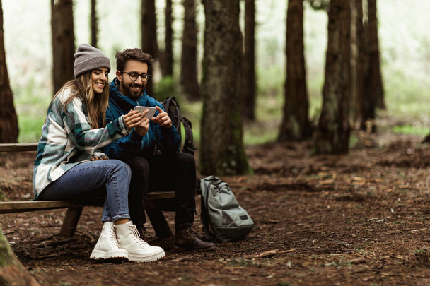Счастливая молодая кавказская семья в куртках отдыхает на скамейке запасных, планирует маршрут на онлайн-карте приложения по телефону, наслаждается приключениями, гуляет в холодном лесу осенью. Фото отдыха вместе, активный образ жизни - Фото, изображение