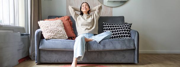 Πορτρέτο της ανέμελης Ασιάτισσας που απολαμβάνει ρεπό, κάθεται στον καναπέ και χαμογελά ευχαριστημένη, χαλαρώνοντας στο σπίτι τα Σαββατοκύριακα. - Φωτογραφία, εικόνα