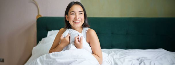 Κοντινό πλάνο της χαρούμενης όμορφης Ασιάτισσας γυναίκας, που ξυπνάει στο κρεβάτι και απολαμβάνει το πρωί, κοιτάζοντας έξω από το παράθυρο με νυσταγμένο χαμόγελο στο πρόσωπό της, αγκαλιάζοντας πάπλωμα. - Φωτογραφία, εικόνα