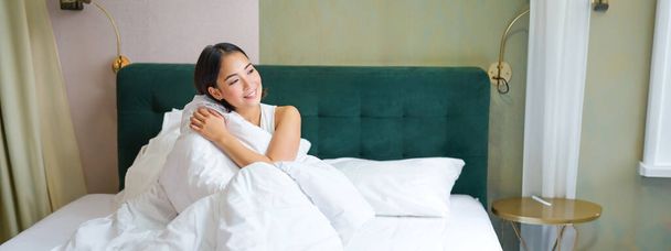 Schöne verschlafene asiatische Mädchen wacht in ihrem Bett mit gemütlichen weißen Decken auf und schaut aus dem Fenster mit zufriedenem Lächeln. Lifestyle-Konzept. - Foto, Bild