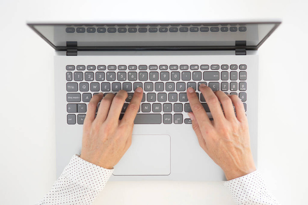 Χέρια μιας γυναίκας στα σαράντα της φορώντας ένα λευκό πουκάμισο φόρεμα με μαύρες κουκίδες, πληκτρολογώντας σε ένα πληκτρολόγιο laptop και σε ένα λευκό τραπέζι. - Φωτογραφία, εικόνα