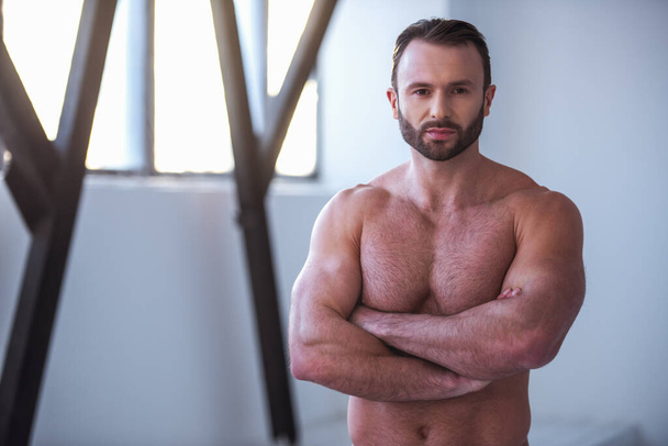 Hombre musculoso guapo con torso desnudo está mirando a la cámara mientras está de pie con los brazos cruzados en el gimnasio
 - Foto, Imagen