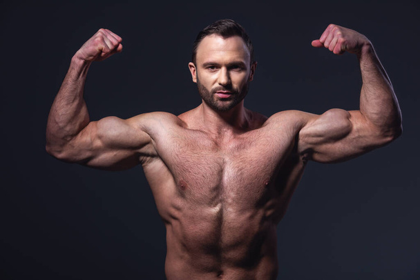 Handsome muscular man with bare torso showing muscles while posing at camera, on dark background - Photo, Image
