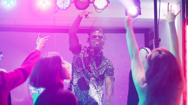 Afrikkalainen amerikkalainen DJ sekoittaen ääntä lavalla yökerhossa, siisti mies juhlimassa ihmisten kanssa tanssilattialla. Funky aikuinen luoda sähköisen sekoittamisen ääniä paneeli asemalla, klubit. Kolmijalka laukaus. - Valokuva, kuva