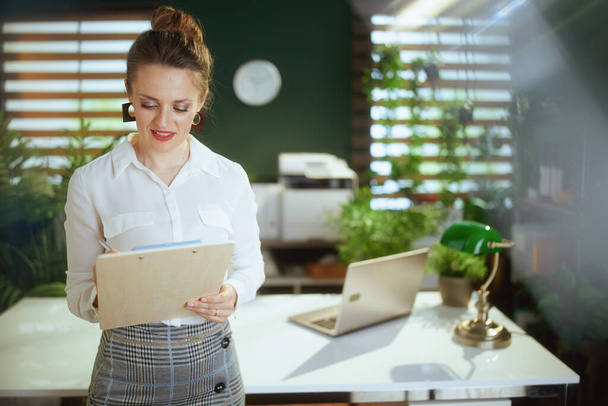 Zrównoważony handel nieruchomościami. zamyślona młoda kobieta agent nieruchomości w nowoczesnym zielonym biurze w białej bluzce z schowkiem. - Zdjęcie, obraz