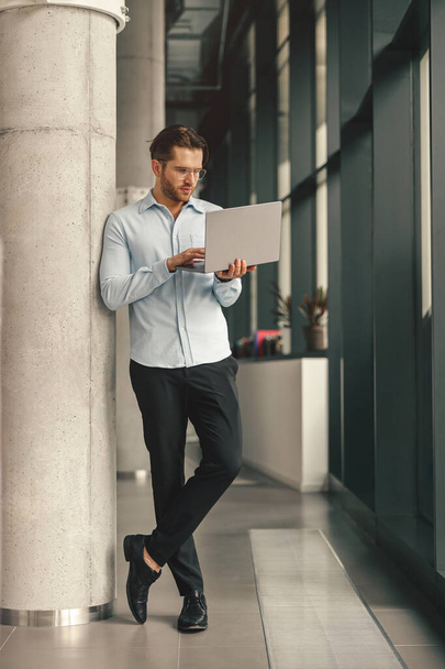 Hezký podnikatel pracující na notebooku stojící v moderní spolupráci během pracovního dne - Fotografie, Obrázek