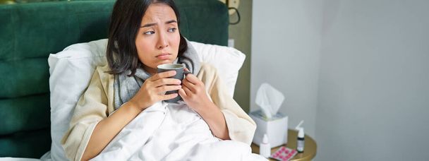 Covid-19, flu and vaccination concept. Korean girl lying in bed with cold, catching influenza, drinking hot tea and using prescribed medication. - Photo, Image