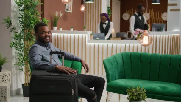 Afro-Amerikalı konuk lobide oturuyor, otele giriş yapmaya hazırlanıyor ve otel hizmetlerinin tadını çıkarıyor, uzun bir uçuştan sonra resepsiyonda dinleniyor. Tatil ve turizm alanlarında dinlenen kişi. - Video, Çekim